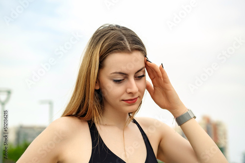 Beautiful brunette woman wearing sports bra standing on city park, outdoors touching her head with hand trying to remember something or having headache. Suffering from pain.