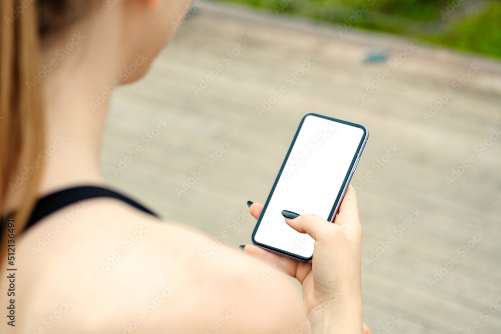 Young brunette woman wearing sportive clothes on city park, outdoors hands holding phone touching finger mockup white blank display, mobile application tech concept, over shoulder closeup view.