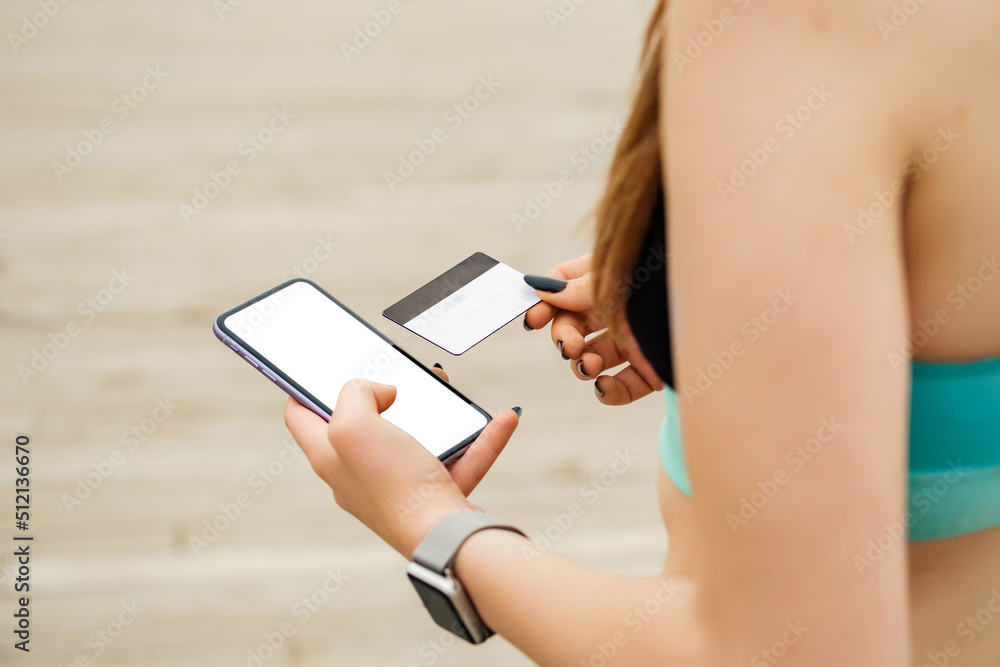 Happy sportive woman standing on city park, outdoors hands holding credit card and phone with mockup white blank display, empty screen for shopping or banking app. Over shoulder close up view.