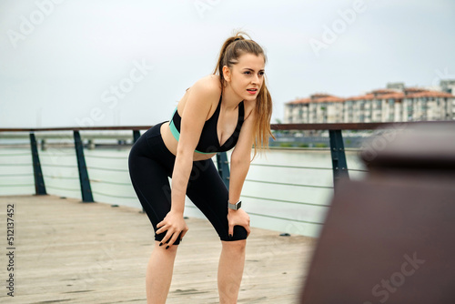 Cute sportive woman wearing black sports bra standing on city park, outdoors standing bent over and catching her breath after a running session. Outdoor sport concepts.