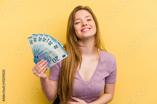 Young caucasian woman holding banknotes isolated on yellow background laughing and having fun.