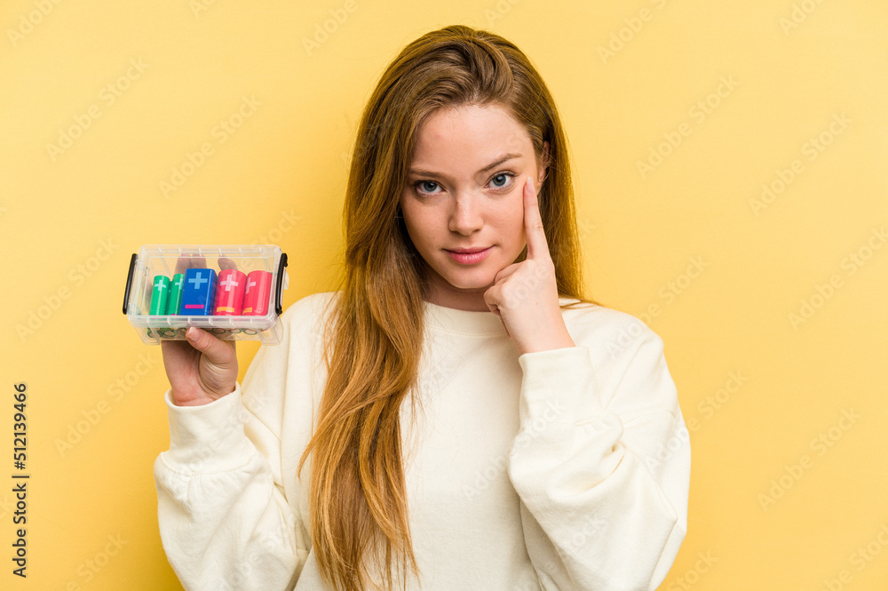 Young caucasian woman holding a battery box isolated on yellow background pointing temple with finger, thinking, focused on a task. Young caucasian woman holding a battery box isolated on yellow background pointing temple with finger, thinking, focused on a task.