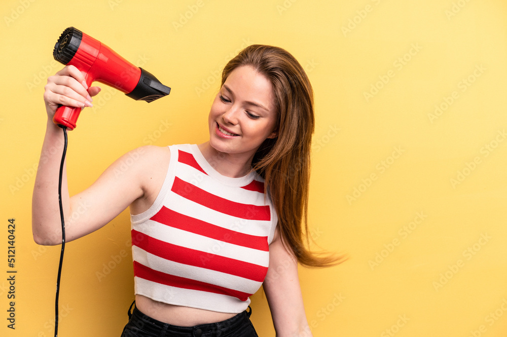 Young caucasian woman holding a hairdryer isolated on yellow background Young caucasian woman holding a hairdryer isolated on yellow background