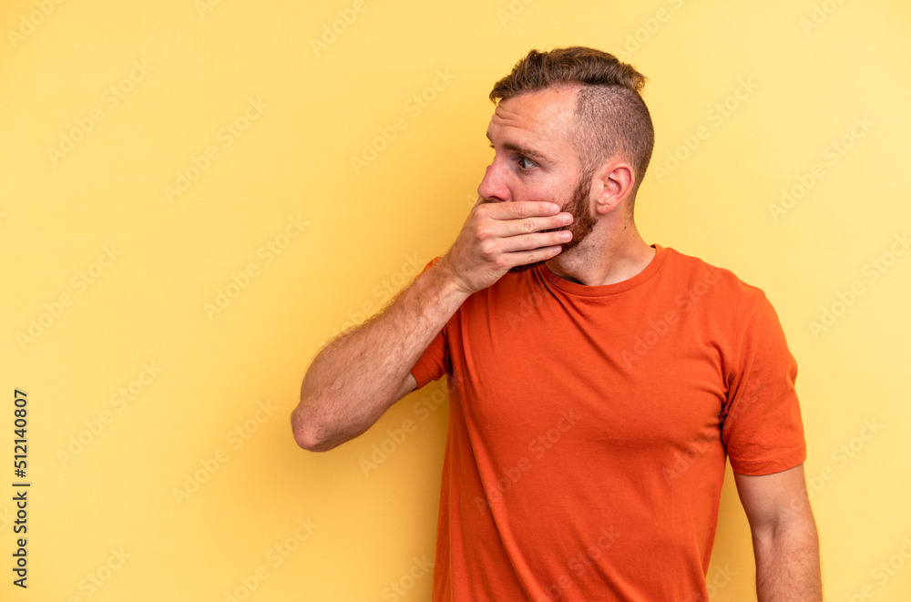 Young caucasian man isolated on yellow background thoughtful looking to a copy space covering mouth with hand. Young caucasian man isolated on yellow background thoughtful looking to a copy space covering mouth with hand.