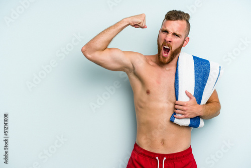 Young caucasian man going to the beach holding a towel isolated on blue background raising fist after a victory, winner concept.