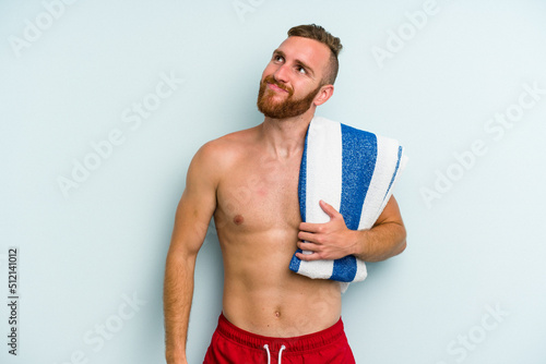 Young caucasian man going to the beach holding a towel isolated on blue background dreaming of achieving goals and purposes
