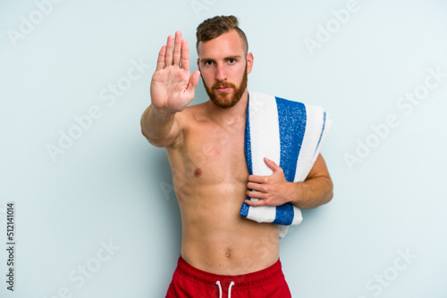 Young caucasian man going to the beach holding a towel isolated on blue background standing with outstretched hand showing stop sign, preventing you.