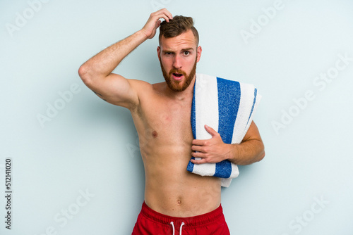 Young caucasian man going to the beach holding a towel isolated on blue background being shocked, she has remembered important meeting.