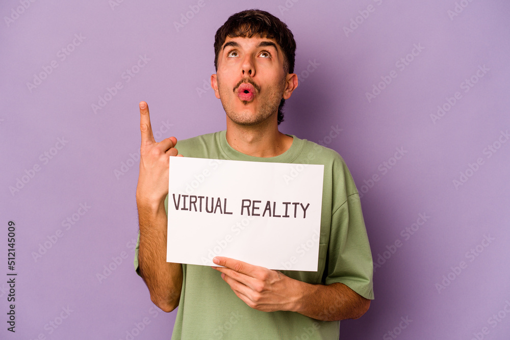 Young hispanic man holding virtual reality placard isolated on purple background pointing upside with opened mouth. Young hispanic man holding virtual reality placard isolated on purple background pointing upside with opened mouth.