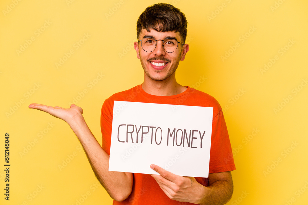 Young hispanic man holding crypto money placard isolated on yellow background showing a copy space on a palm and holding another hand on waist. Young hispanic man holding crypto money placard isolated on yellow background showing a copy space on a palm and holding another hand on waist.