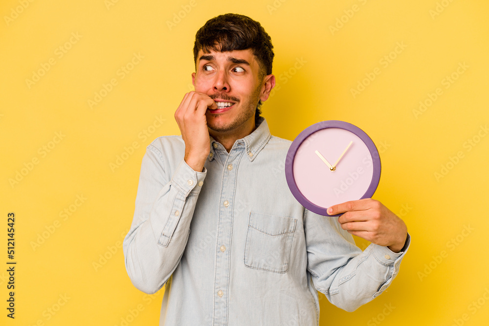Young hispanic man holding a clock isolated on yellow background biting fingernails, nervous and very anxious. Young hispanic man holding a clock isolated on yellow background biting fingernails, nervous and very anxious.