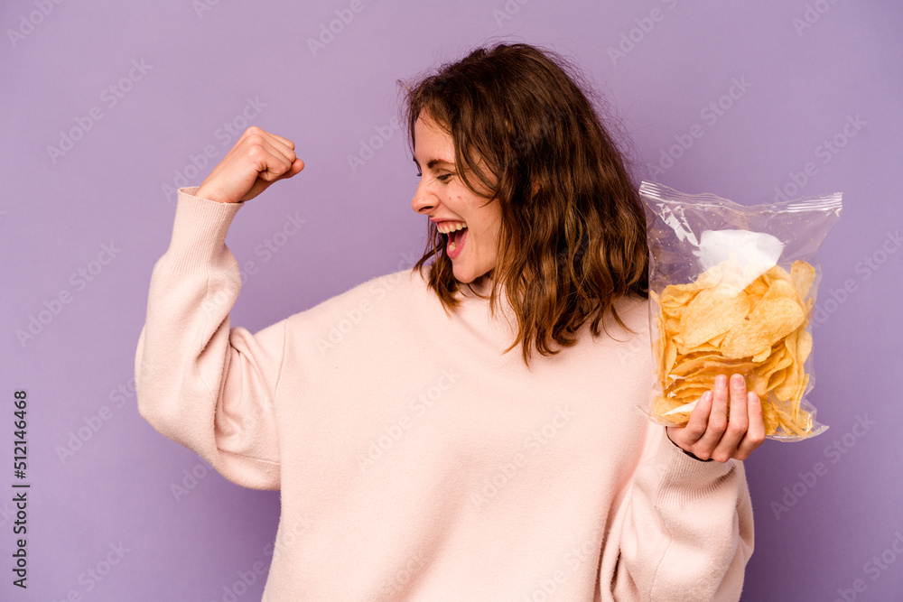 Young caucasian woman holding a bag of chips isolated on purple background raising fist after a victory, winner concept. Young caucasian woman holding a bag of chips isolated on purple background raising fist after a victory, winner concept.