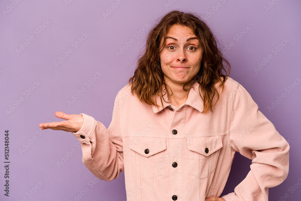 Young caucasian woman isolated on purple background showing a copy space on a palm and holding another hand on waist. Young caucasian woman isolated on purple background showing a copy space on a palm and holding another hand on waist.