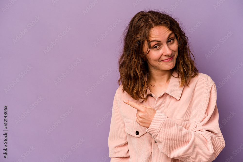 Fototapeta premium Young caucasian woman isolated on purple background smiling and pointing aside, showing something at blank space.
