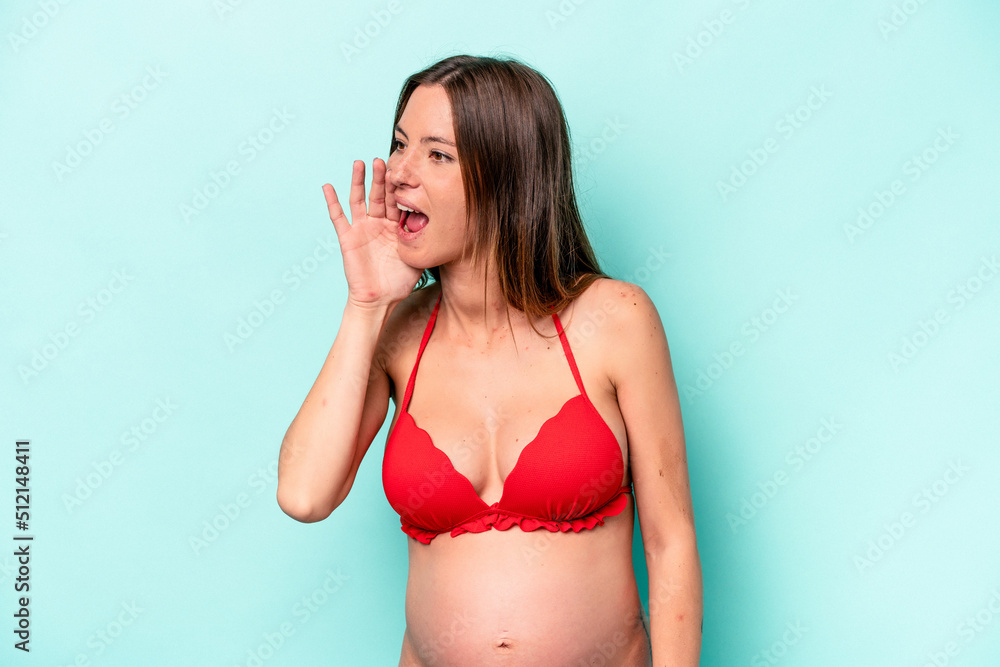 Fototapeta premium Young caucasian pregnant woman wearing bikini isolated on blue background shouting and holding palm near opened mouth.