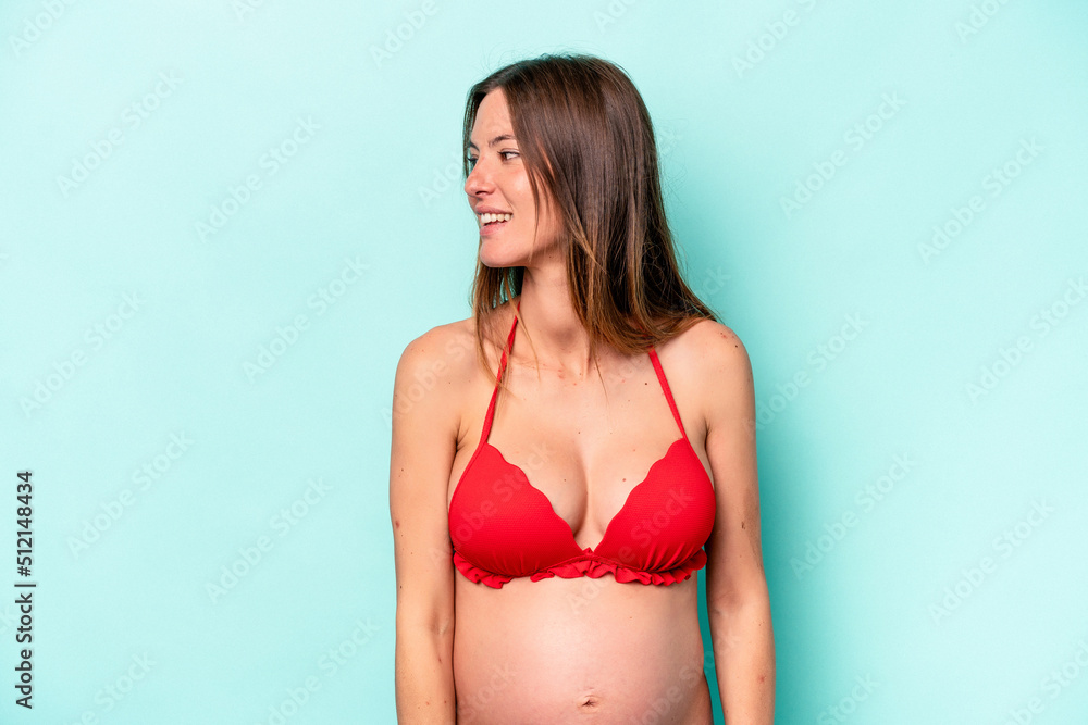 Obraz premium Young caucasian pregnant woman wearing bikini isolated on blue background looks aside smiling, cheerful and pleasant.