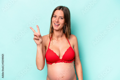Young caucasian pregnant woman wearing bikini isolated on blue background joyful and carefree showing a peace symbol with fingers. © Asier