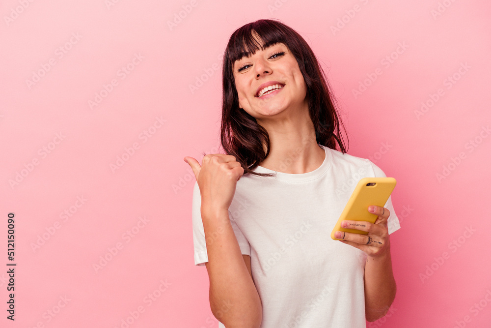 Young caucasian woman holding a mobile phone isolated on pink background points with thumb finger away, laughing and carefree. Young caucasian woman holding a mobile phone isolated on pink background points with thumb finger away, laughing and carefree.
