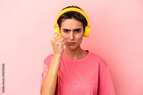 Young caucasian woman listening to music isolated on pink background showing fist to camera, aggressive facial expression.