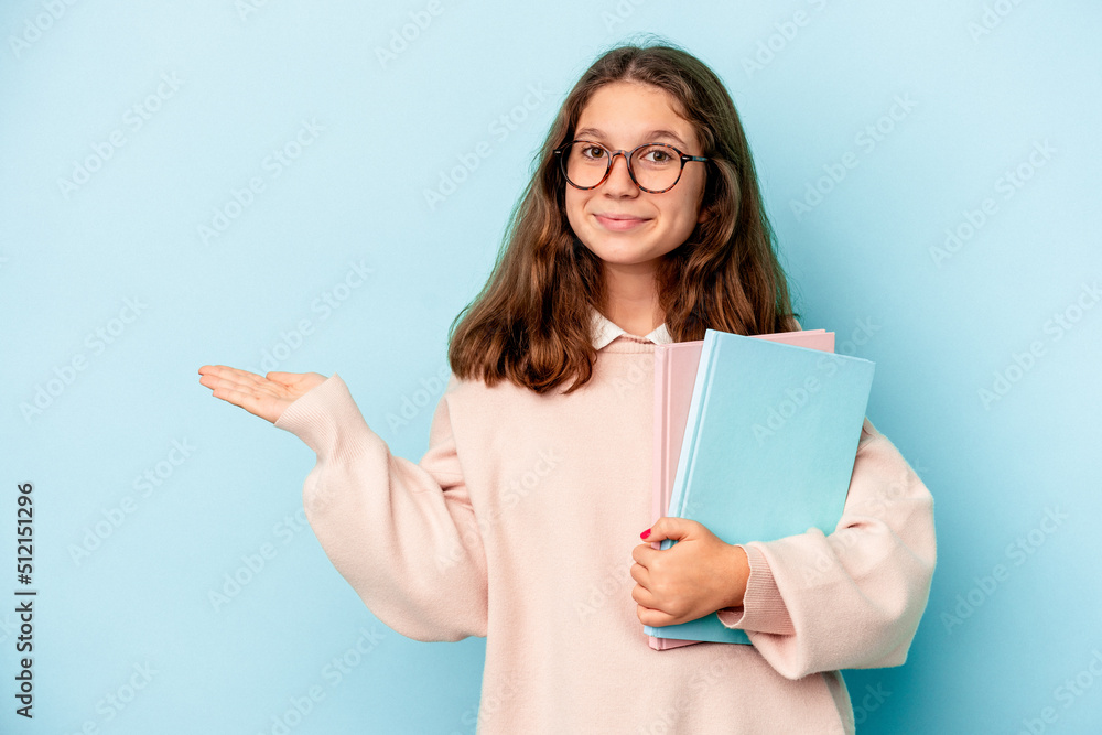 Little caucasian student girl holding books isolated on blue background showing a copy space on a palm and holding another hand on waist. Little caucasian student girl holding books isolated on blue background showing a copy space on a palm and holding another hand on waist.