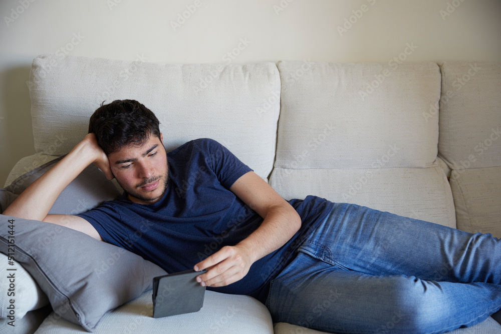 Young man lying down on the sofa reading an ebook