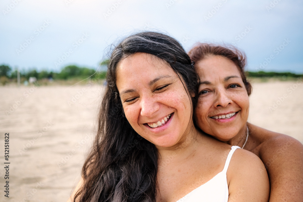 close up of old mother and pregnant adult daughter are seen hugging and smiling on a beach with white dress close up of old mother and pregnant adult daughter are seen hugging and smiling on a beach with white dress