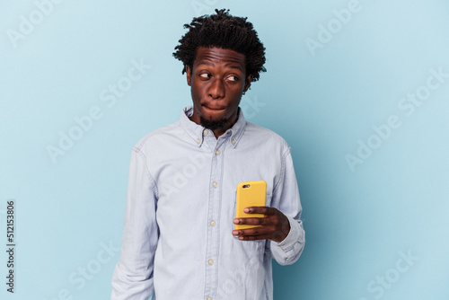 Young african american man holding mobile phone isolated on blue background confused  feels doubtful and unsure.