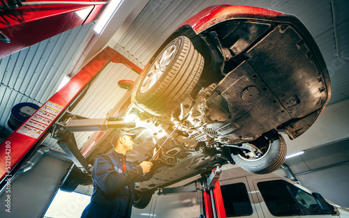 Young car mechanic at repair service station inspecting car wheel and suspension detail of lifted automobile. Bottom view.