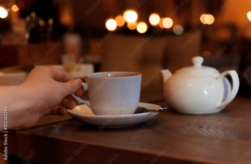 White cup with teapot. The hand holding the cup of tea standing on saucer in soft focus on naturally blurred background. Coffee, tea house, bokeh lights. The concept of a cozy pastime White cup with teapot. The hand holding the cup of tea standing on saucer in soft focus on naturally blurred background. Coffee, tea house, bokeh lights. The concept of a cozy pastime