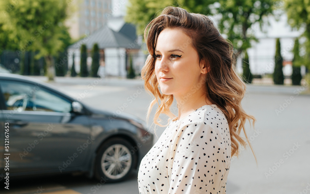 Stylish young woman with curly hairstyle standing near road and car in city. Portrait of cute pretty lady model looking away on street, side view Stylish young woman with curly hairstyle standing near road and car in city. Portrait of cute pretty lady model looking away on street, side view