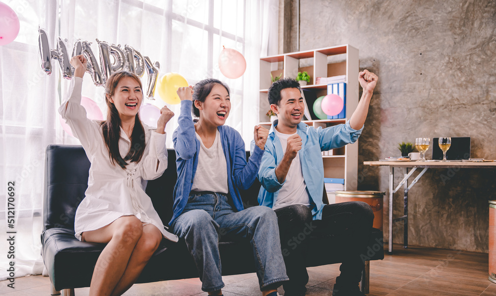 A group of Asian friends sit at home watching on the sofa and cheering for a sporting match on TV together. Excited male and female sports fans celebrate team sports victories at national games. A group of Asian friends sit at home watching on the sofa and cheering for a sporting match on TV together. Excited male and female sports fans celebrate team sports victories at national games.