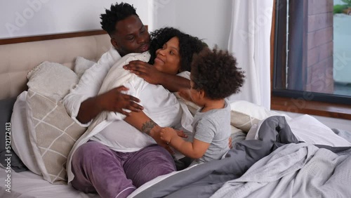 Loving African American young husband hugging pregnant wife talking to little cute son. Happy man enjoying weekend leisure with family in bedroom at home. Relaxation and care concept photo