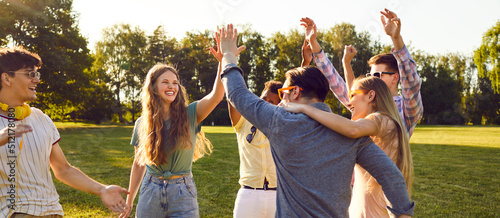 Bunch of happy young friends all together having fun on warm sunny day in summer park. Diverse group of cheerful joyful positive people standing on green lawn, smiling and giving each other high five photo