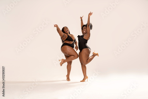 Full photo of superb plus size dark skinned two women in black fashionable swimwears, laughing and dancing, having fun together. Concept of body acceptance, body positivity