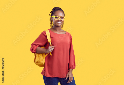 Portrait of young african american woman with backpack on shoulders isolated on yellow background. Smiling student girl in bright glasses and casual clothes looks at camera. Educational banner.
