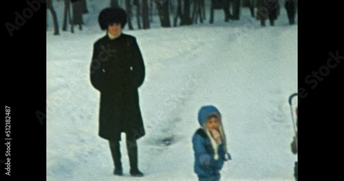 Small child stands in snow winter park. Mother follows baby on family walk in forest. Funny calm little kid in cold outdoor dressed in blue jumpsuit jacket. Vintage color film. Retro archive 1980s