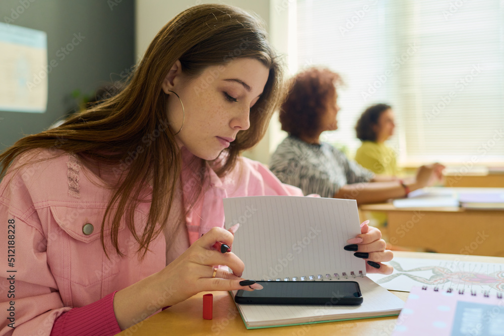 Teenage girl in pink denim jacket texting in smartphone at lesson behind page of copybook while sitting by desk against her classmates Teenage girl in pink denim jacket texting in smartphone at lesson behind page of copybook while sitting by desk against her classmates