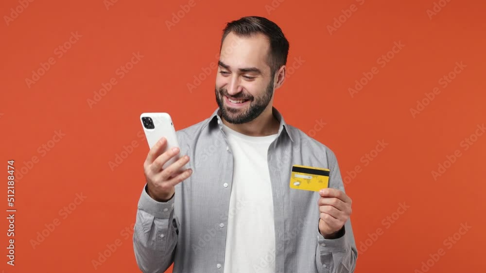 Young brunet man 20s wear blue shirt using mobile cell phone hold in hand credit bank card doing online shopping order delivery to home booking tour isolated on plain orange background studio portrait Young brunet man 20s wear blue shirt using mobile cell phone hold in hand credit bank card doing online shopping order delivery to home booking tour isolated on plain orange background studio portrait