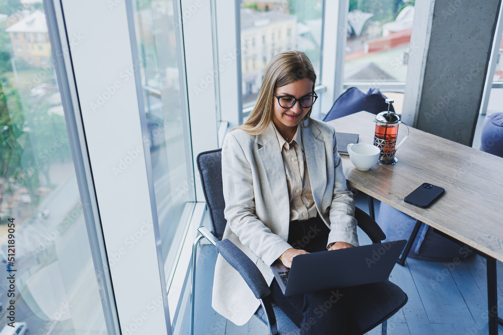 Smiling business woman looking at laptop while working in office. The concept of a modern successful woman. The idea of business and life of an entrepreneur. Young woman at table with open laptop Smiling business woman looking at laptop while working in office. The concept of a modern successful woman. The idea of business and life of an entrepreneur. Young woman at table with open laptop