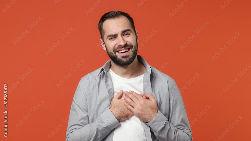 Blithesome good kind fun young brunet man 20s years old wears blue shirt looking camera put folded hands on heartisolated on plain orange background studio portrait. People emotions lifestyle concept Blithesome good kind fun young brunet man 20s years old wears blue shirt looking camera put folded hands on heartisolated on plain orange background studio portrait. People emotions lifestyle concept