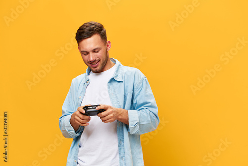 Overjoyed tanned handsome gamer man in blue basic t-shirt hold joystick look at gamepad posing isolated on orange yellow studio background. Copy space Banner Mockup. People lifestyle concept © SHOTPRIME STUDIO