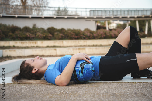 Serious young female football player lying on ground in park