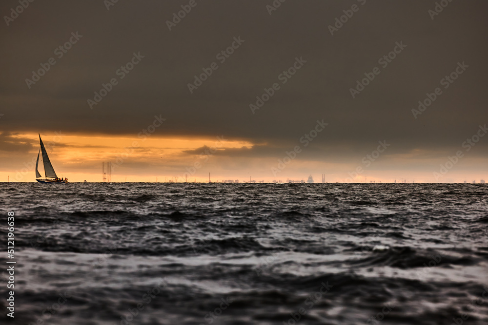 Sailboat in sea at stormy weather, stormy clouds sky orange sky, sail regatta, reflection of sail in the water, bigl waves of water, 