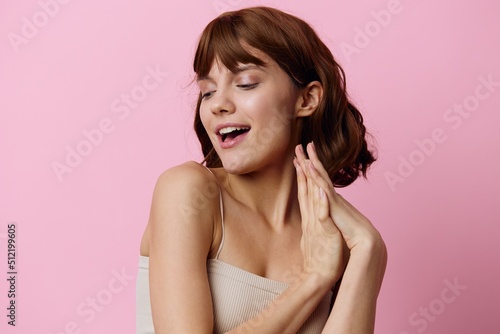 a modest, shy, attractive woman stands on a pink background and smiles pleasantly looking at the camera and holds her hand near her face in a gentle gesture