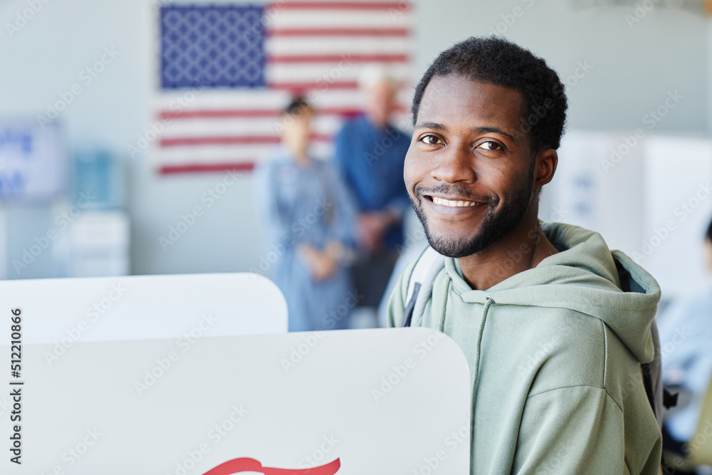 © Seventyfour - Side view portrait of young black man voting in booth on election day and smiling at camera, copy space © Seventyfour - Side view portrait of young black man voting in booth on election day and smiling at camera, copy space