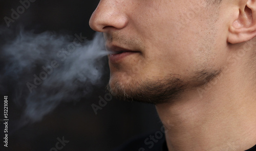 close up of a man with an electronic cigarette. man smokes an electronic cigarette in a natural background. hipster smokes an e-cigarette, breathes out streams of smoke and vaping outdoors copy space