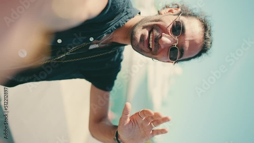 VERTICAL VIDEO: Clouse-up,young italian guy with long curly hair and stubble takes selfie on mobile phone. Stylish man in sunglasses posing smiling at camera of his phone and pointing at yacht  photo