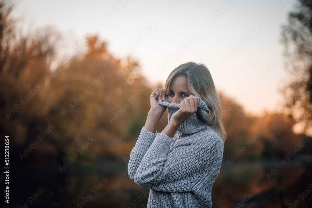 Charming blonde woman standing by river in forest, looking away smiling and pulling sweater collar over face with trees covered with sunset in background. Autumn walk in forest. Charming blonde woman standing by river in forest, looking away smiling and pulling sweater collar over face with trees covered with sunset in background. Autumn walk in forest.