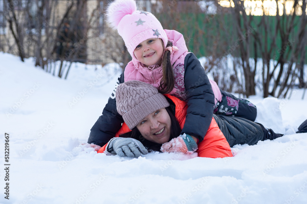 Caucasian happy little daughter with mother lying on snow together smiling in park in evening with trees in background. Parents spending time with children.