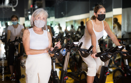 Young girl and her adult mother in face masks training on fitness bikes in gym